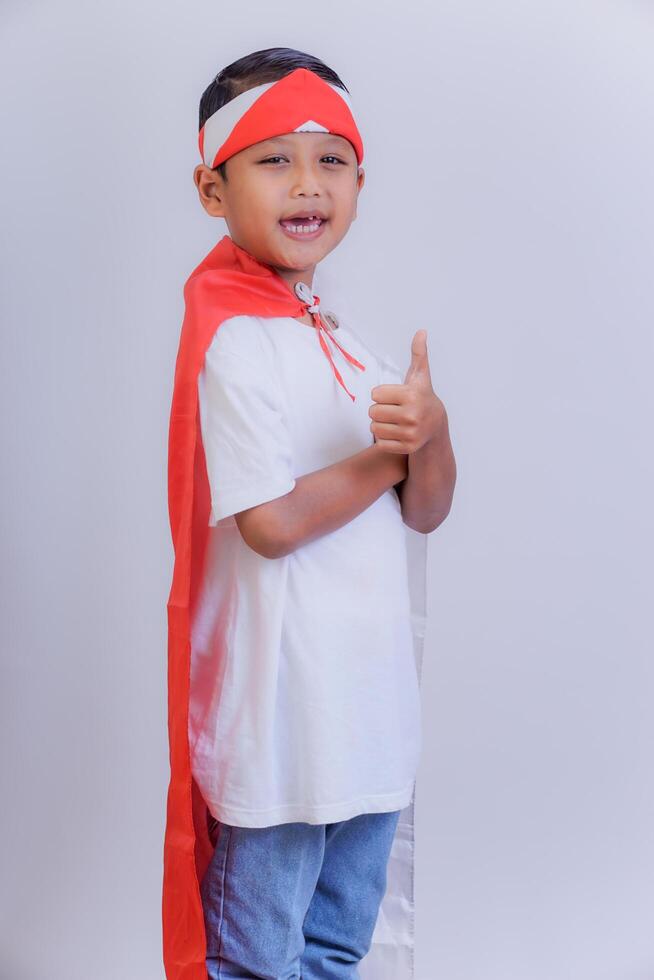 A boy wearing a red and white headband and Indonesian flag on his back cape gives a thumbs-up, smiling confidently. photo