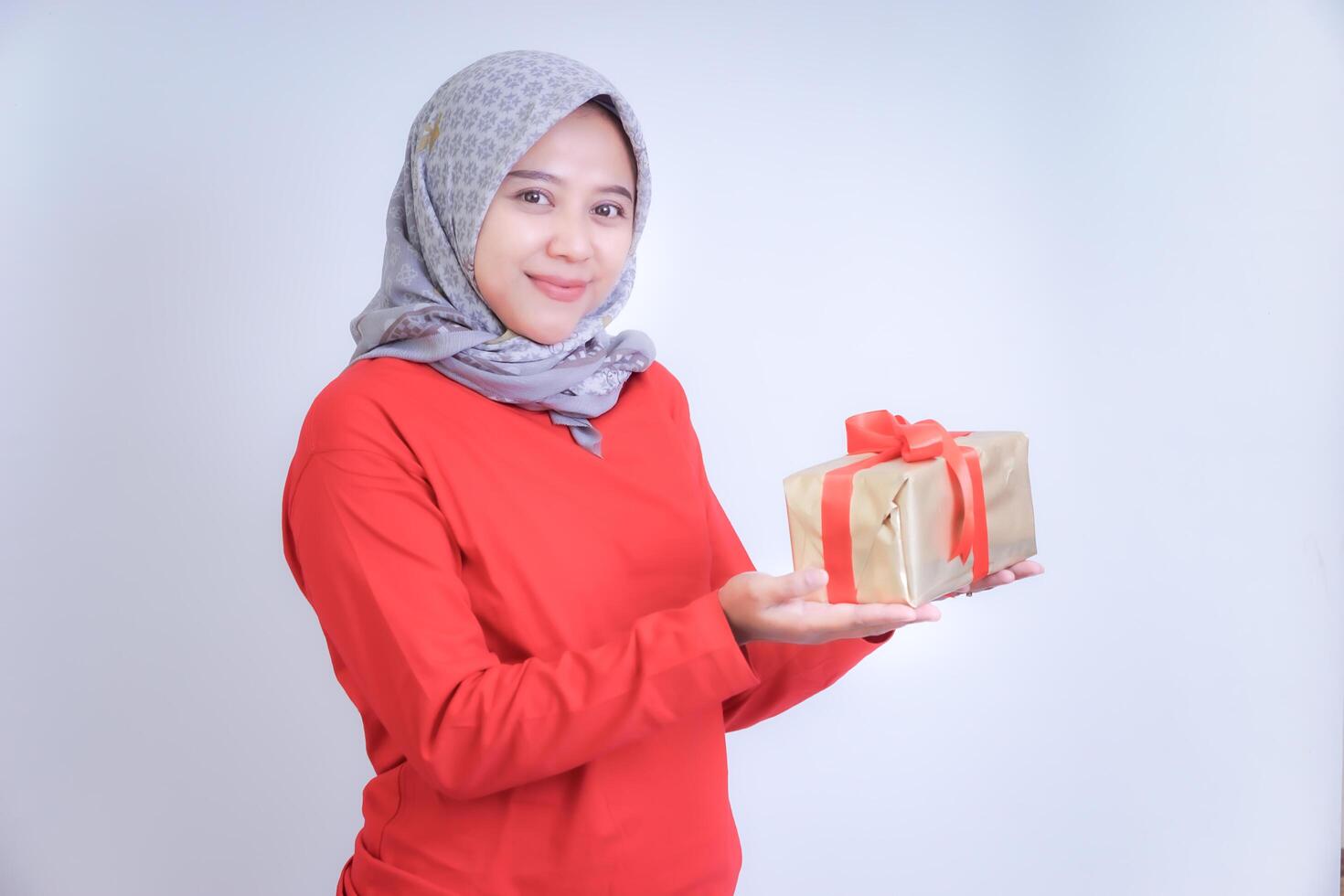 A woman in a red blouse and patterned grey hijab smiles while holding a gift wrapped with a red ribbon. photo