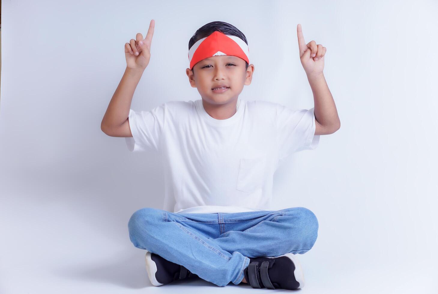 A boy wearing a white shirt and jeans sits cross-legged, pointing up with both hands, smiling brightly. photo