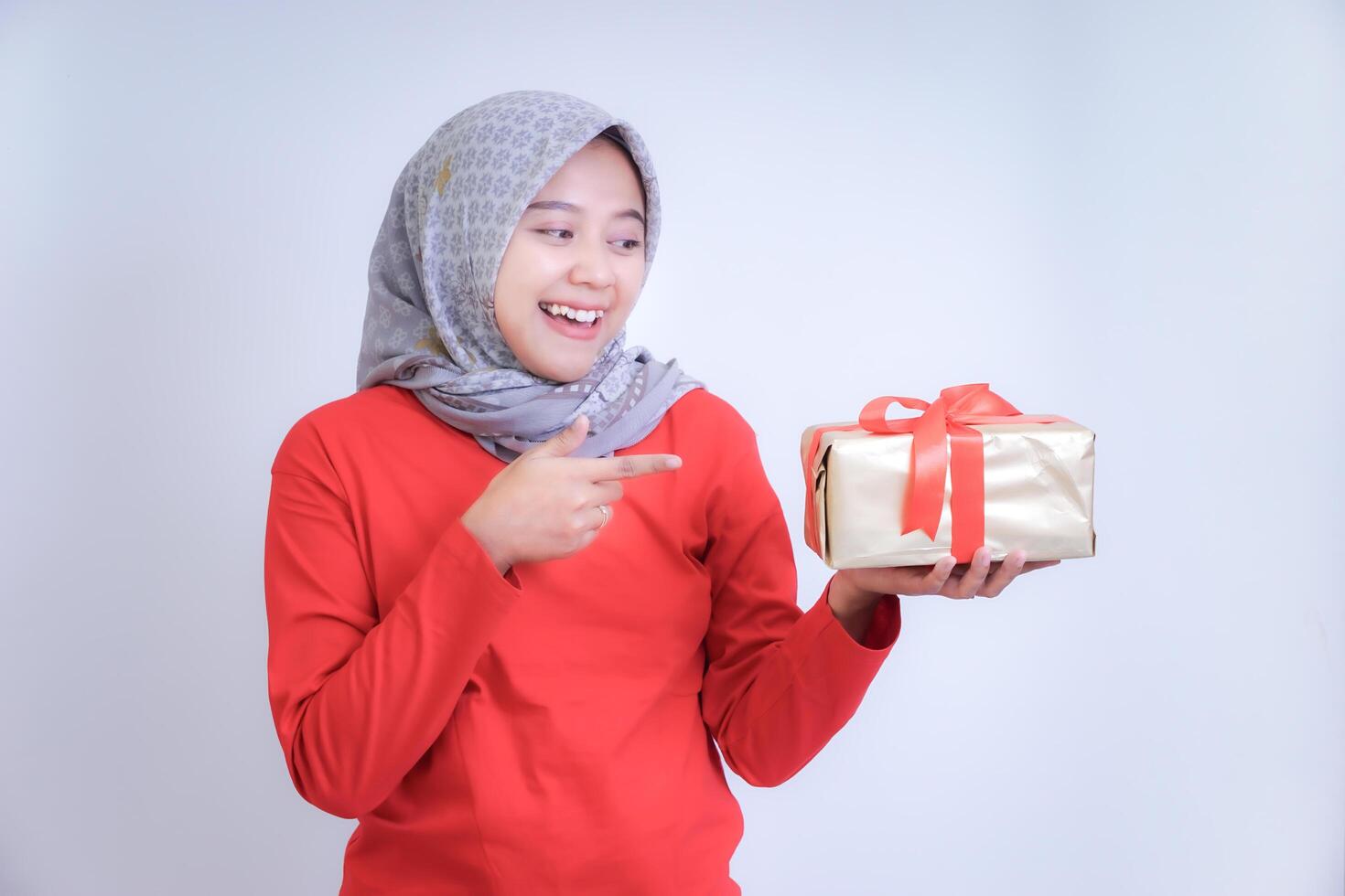 A woman wearing a hijab and red shirt is smiling and holding a gift wrapped in gold paper with a red ribbon. photo