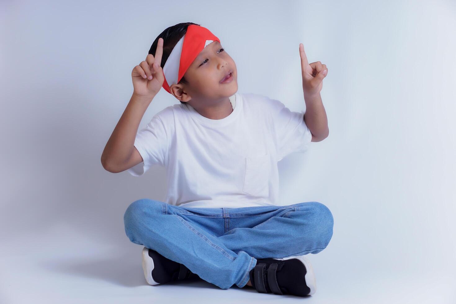 A boy wearing a white shirt and jeans sits cross-legged, pointing up with both hands, smiling brightly. photo