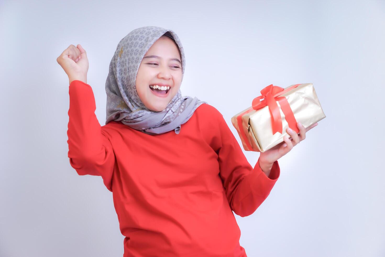 A joyful woman in a red outfit and hijab is celebrating while holding a gift with a red ribbon. photo