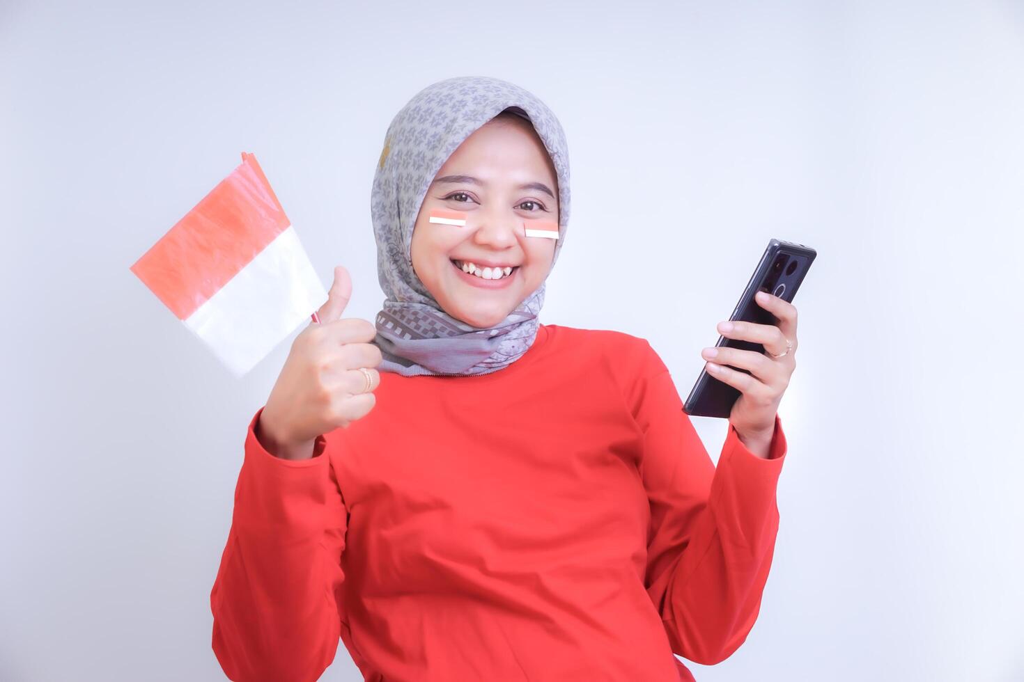 A woman wearing a red outfit smiles while holding an Indonesian flag and a phone, celebrating Indonesia Independence Day. photo