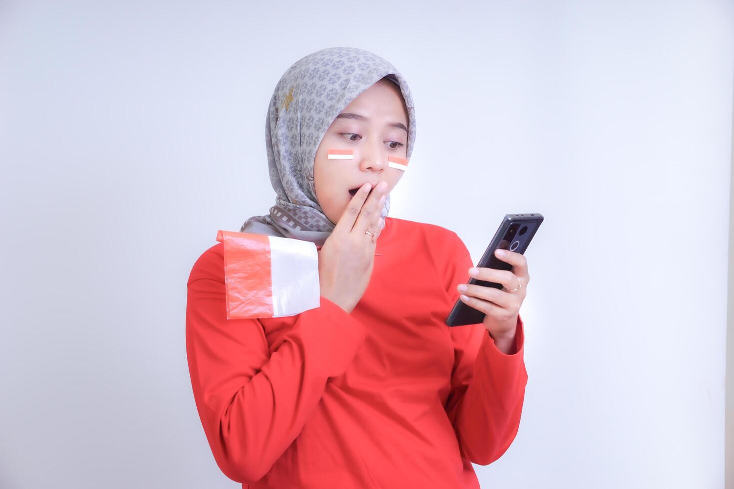 A woman in a red dress with a surprised expression looks at her cell phone while holding an Indonesian flag, celebrating Indonesia's Independence Day. photo