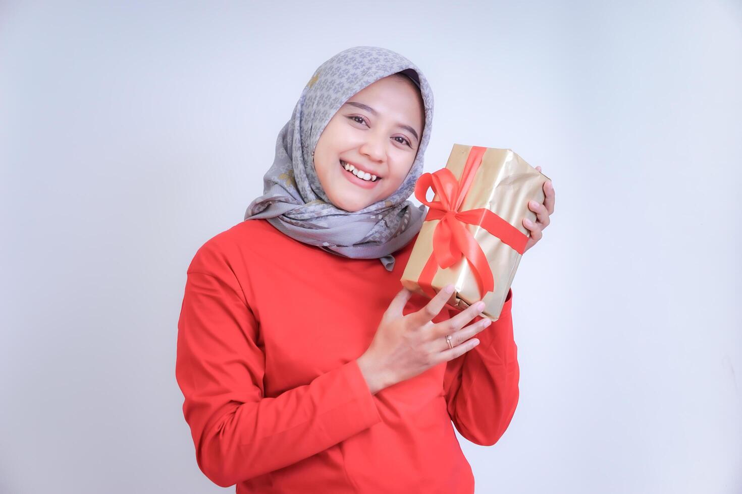 A woman in a red shirt and grey hijab smiles while holding a gift wrapped in gold paper with a red ribbon. photo