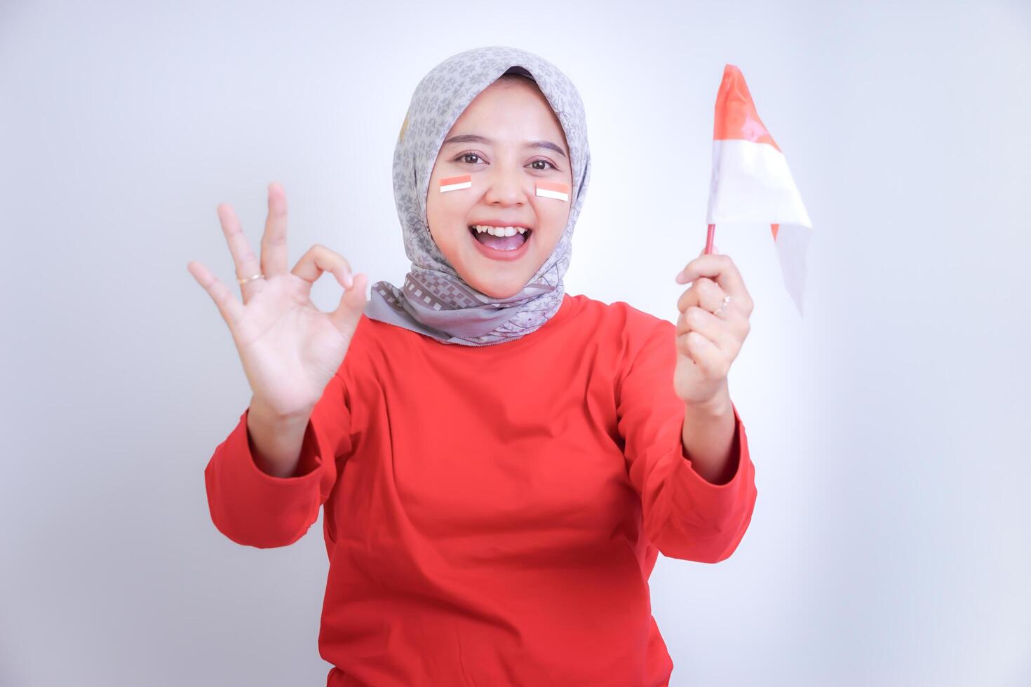 A cheerful woman in a red shirt, holding an Indonesian flag, and making an OK sign, concept of celebrating Indonesia Independence Day. photo
