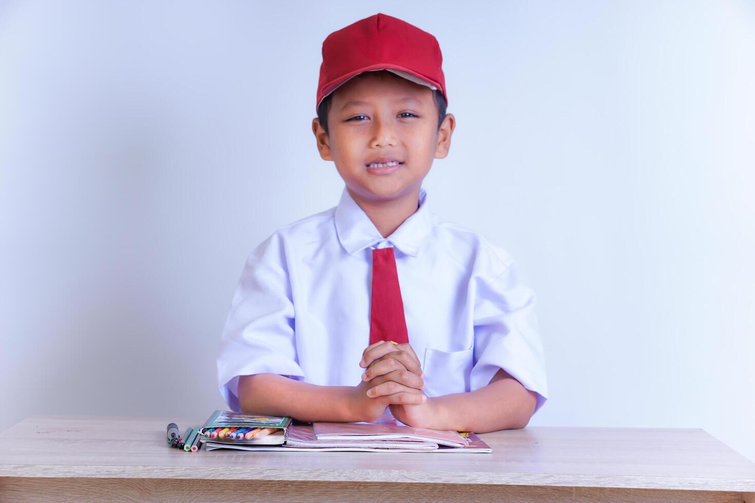 An Indonesian boy in school uniform sits at a table, wearing a white shirt, red tie and red hat. He is smiling with his hands clenched. School supplies sit on the table. The plain background. photo
