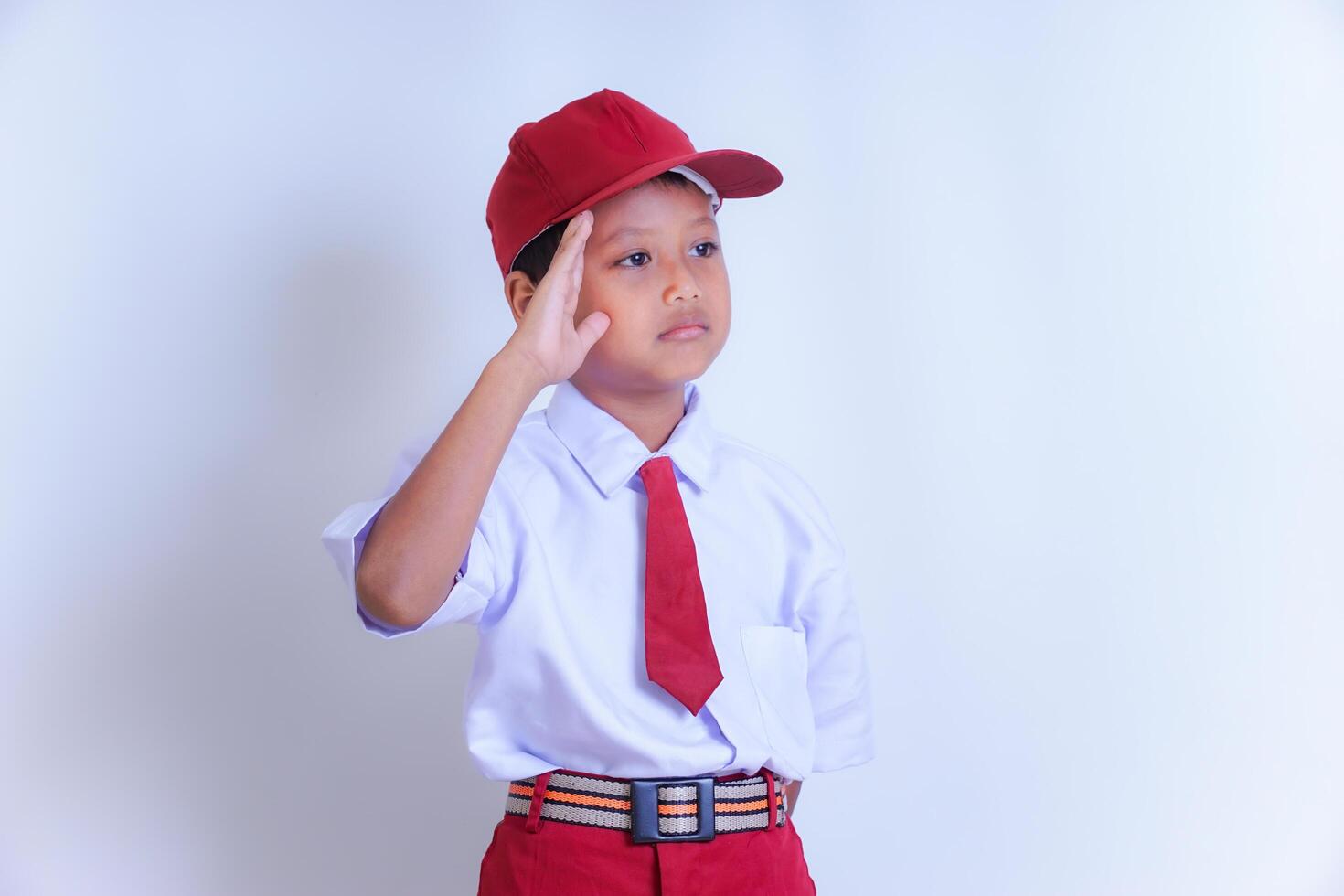 A Indonesian young boy in a school uniform is saluting with his right hand. He wears a white short-sleeved shirt, red tie, red trousers, and a red cap, looking serious and focused. photo