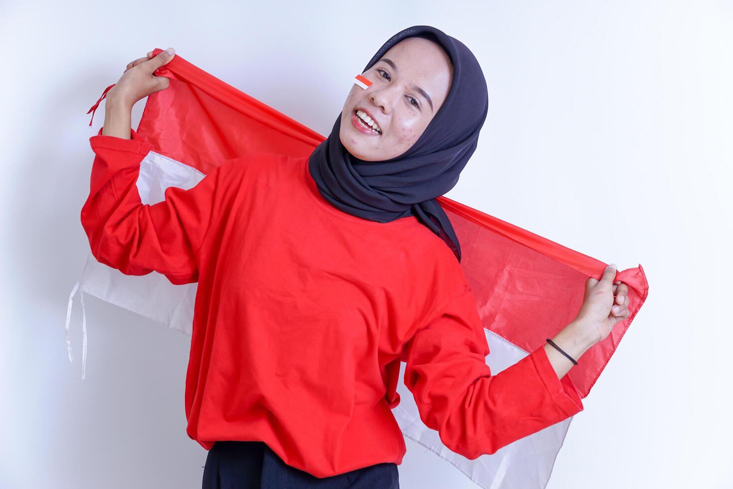 A Indonesian young woman wearing hijab is joyfully holding up the red and white national flag, her face adorned with a smile and a small flag sticker on her cheek, Isolated on white background photo