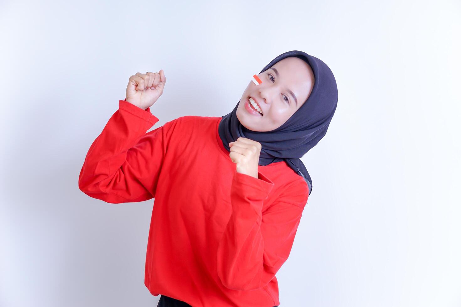 A young Indonesian woman wearing a hijab is smiling happily, wearing a red T-shirt and has a small flag sticker on her cheek, signifying a celebration or national pride. photo