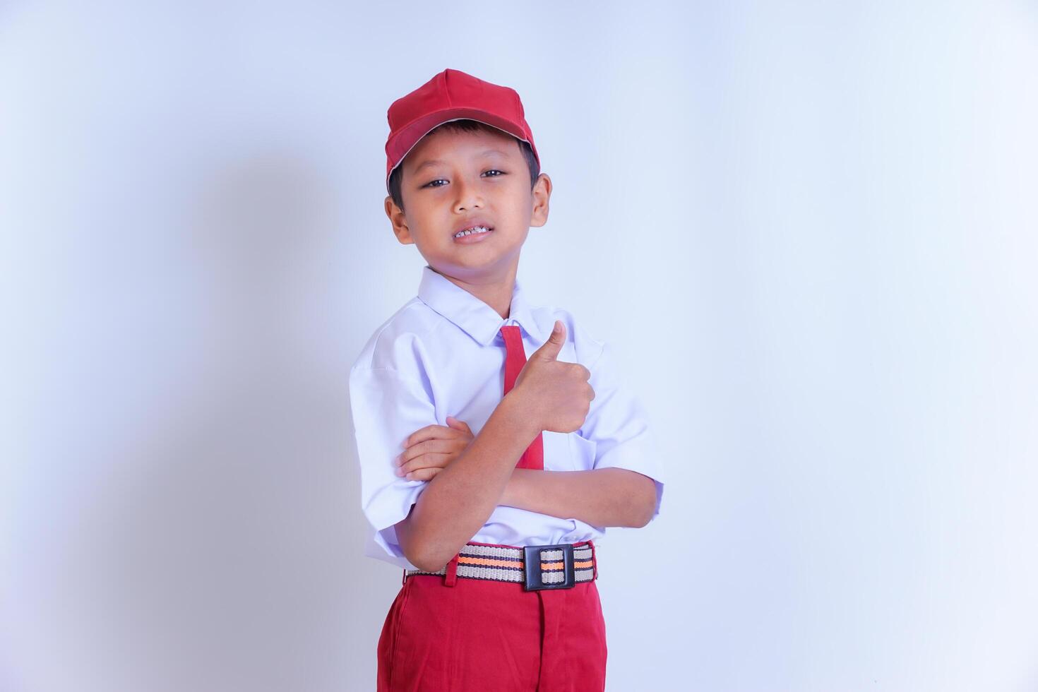 An Indonesian boy in school uniform stands confidently with a thumbs-up, wearing a red cap, white shirt, red shorts and a striped belt. photo