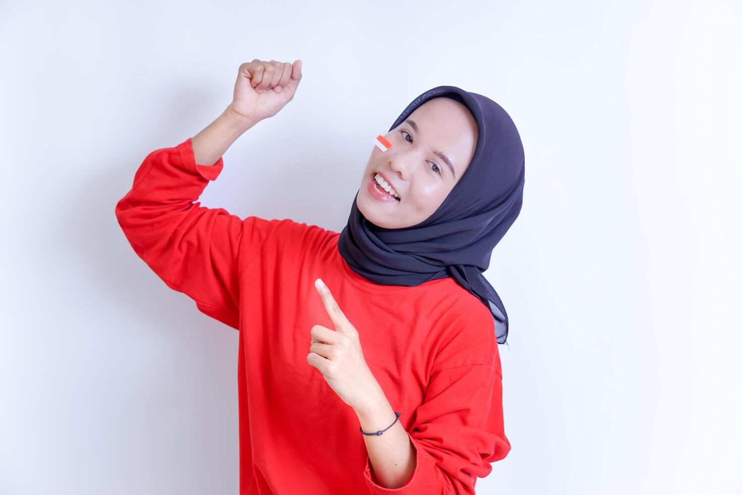 A young Indonesian woman wearing a hijab poses with a cheerful expression, her right hand clenched into a fist and her left hand pointing upwards. photo