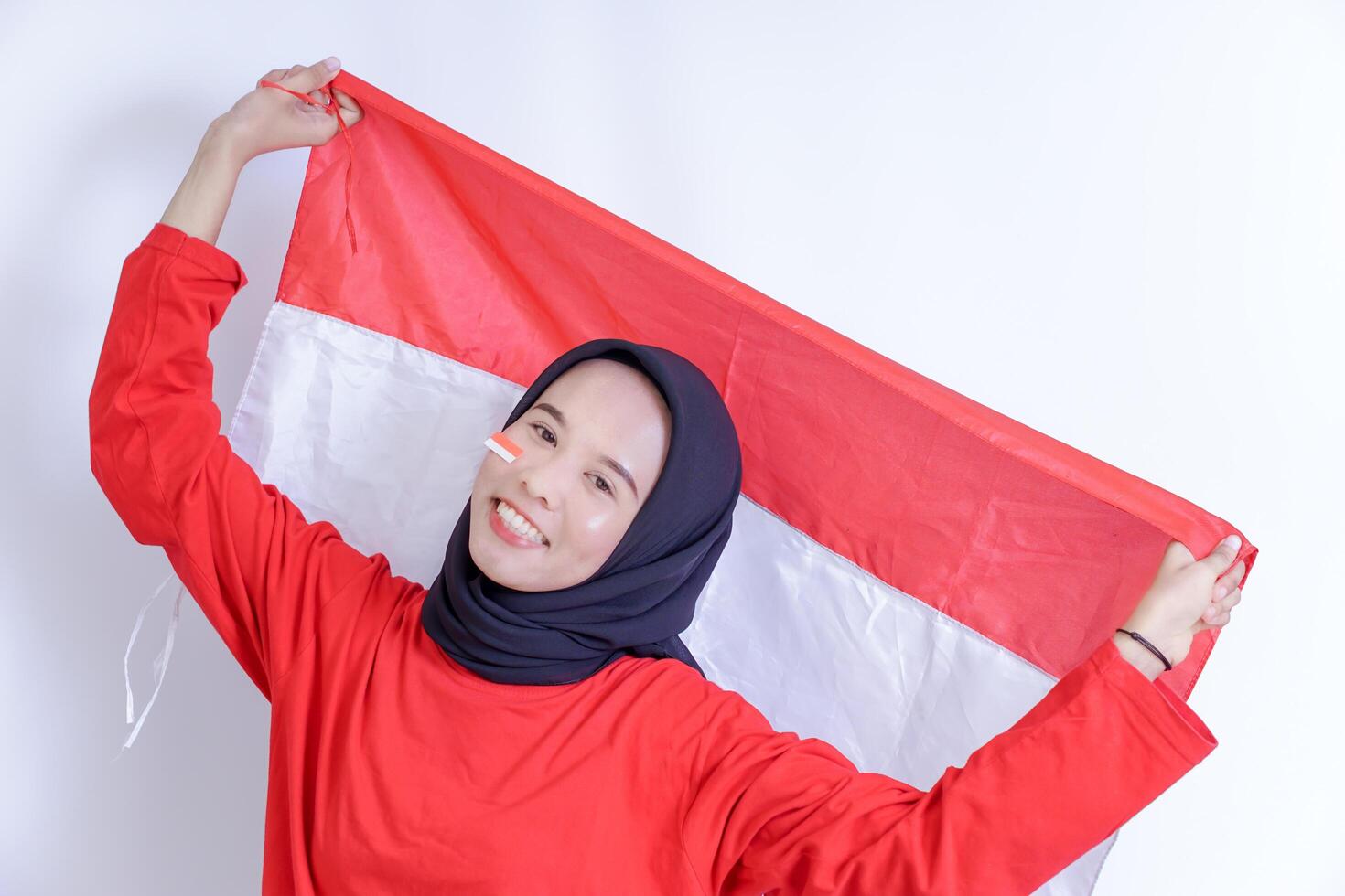 A Indonesian young woman wearing hijab is joyfully holding up the red and white national flag, her face adorned with a smile and a small flag sticker on her cheek, Isolated on white background photo