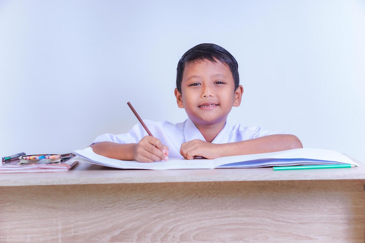 A Indonesian young boy in a school uniform is studying at a desk with pencils and books. photo