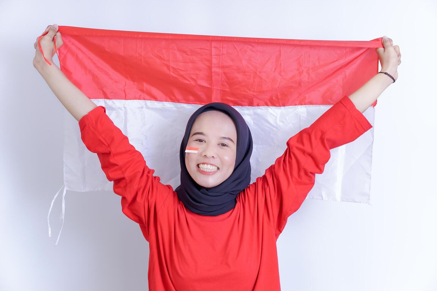 A Indonesian young woman wearing hijab is joyfully holding up the red and white national flag, her face adorned with a smile and a small flag sticker on her cheek, Isolated on white background photo