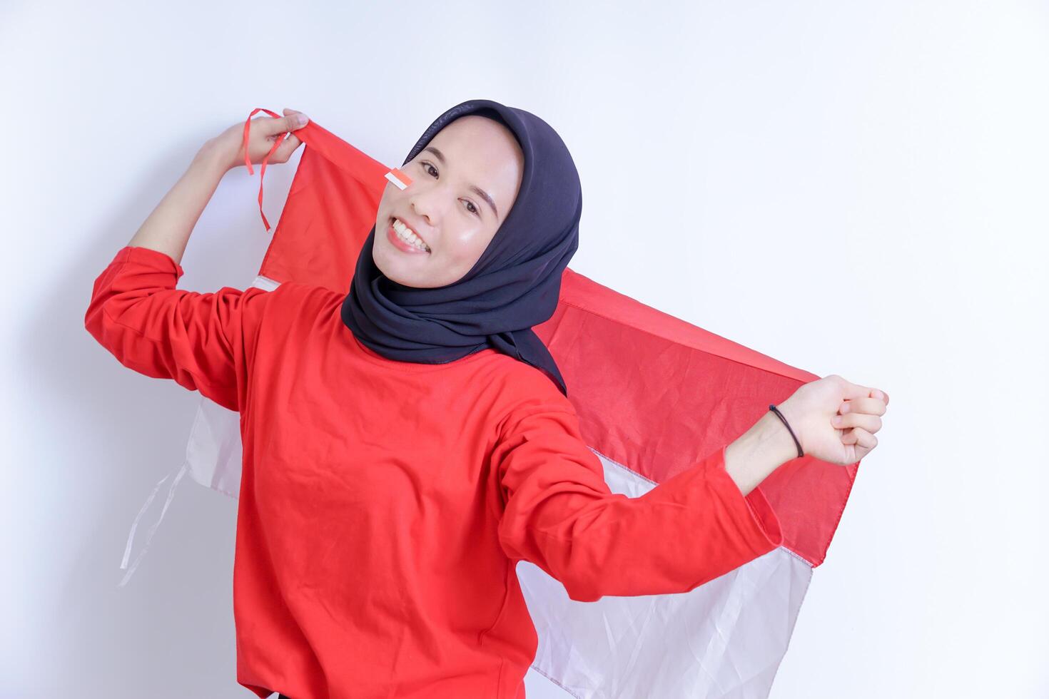 A Indonesian young woman wearing hijab is joyfully holding up the red and white national flag, her face adorned with a smile and a small flag sticker on her cheek, Isolated on white background photo