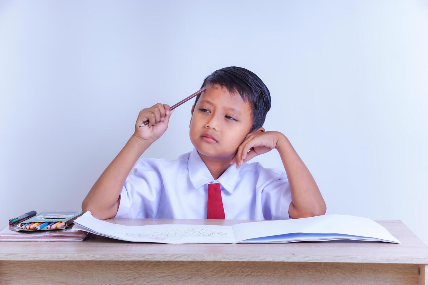 A Indonesian young boy in a school uniform looks thoughtful with a pencil to his head, sitting at a desk with an open notebook and colors. photo