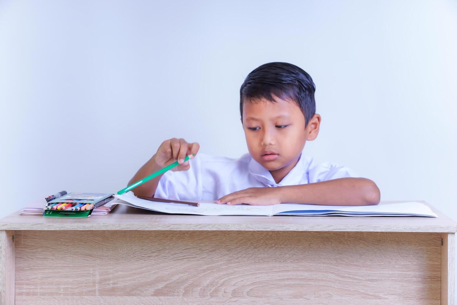A Indonesian young boy in a school uniform is studying at a desk with pencils and books. photo