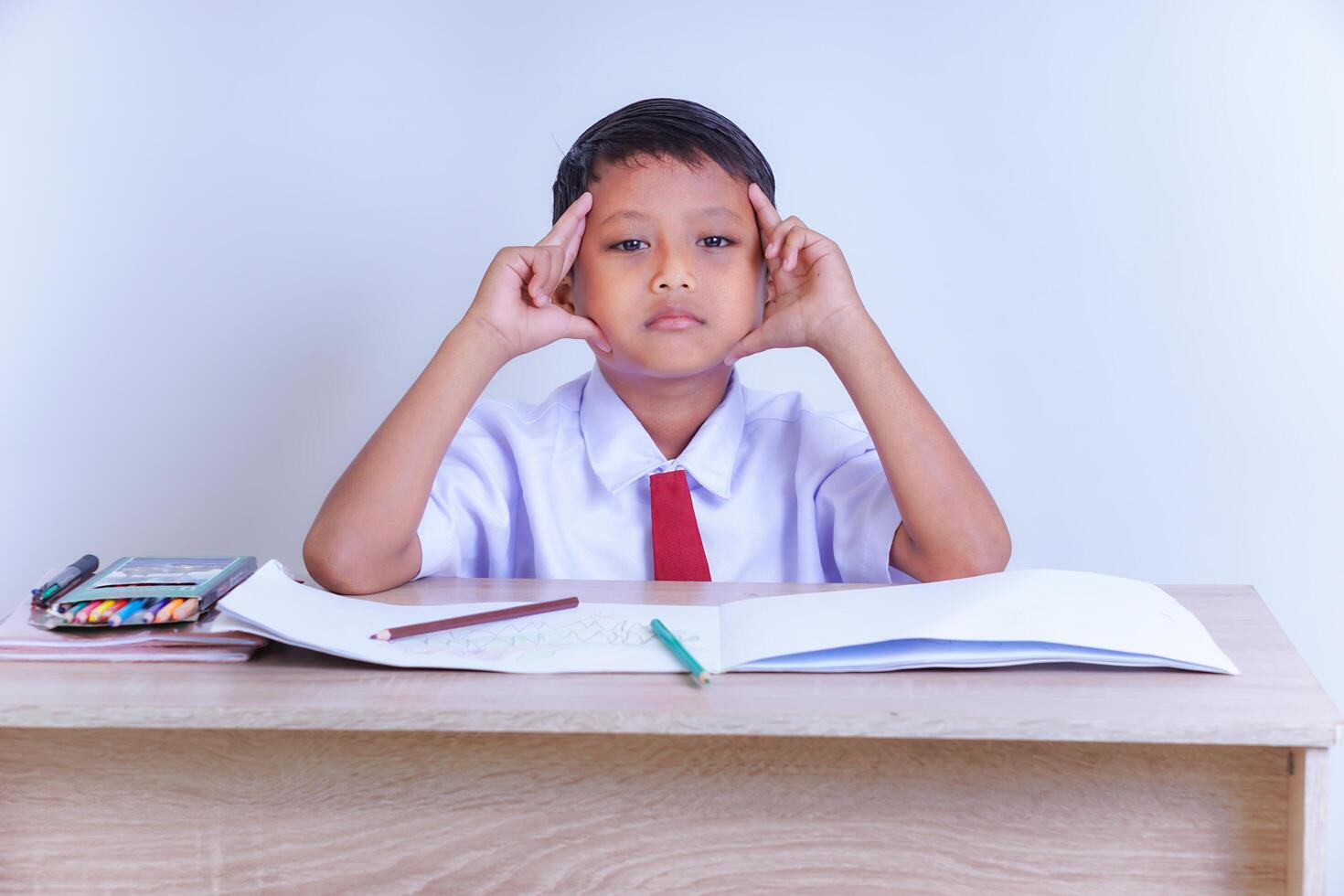 A Indonesian young boy in a school uniform sits at a desk, looking tired or bored, with his hands on his head and school supplies nearby. photo