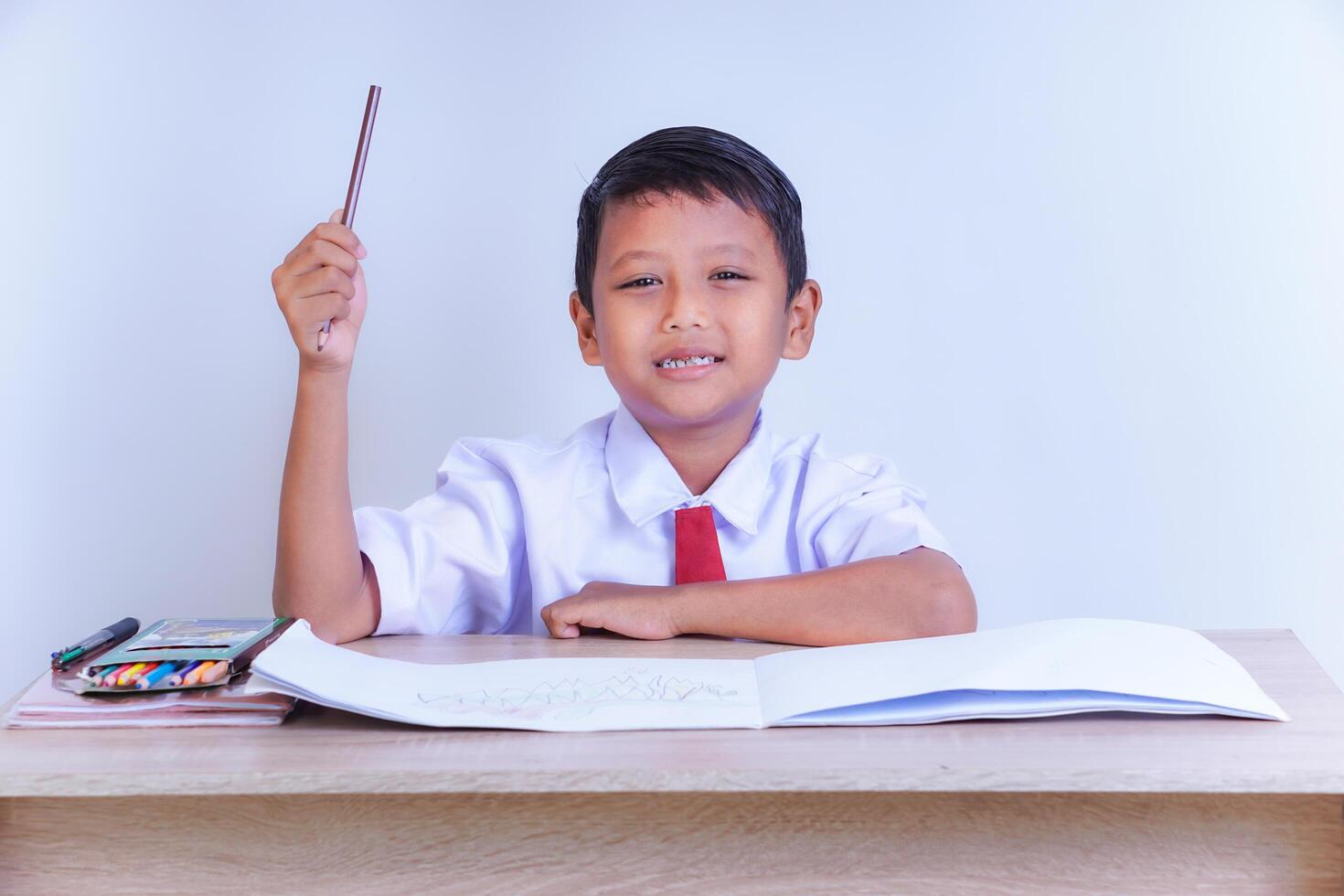 A Indonesian young boy in a school uniform, sitting at a desk with a raised hand, smiling, with books and crayons, ready to learn. photo