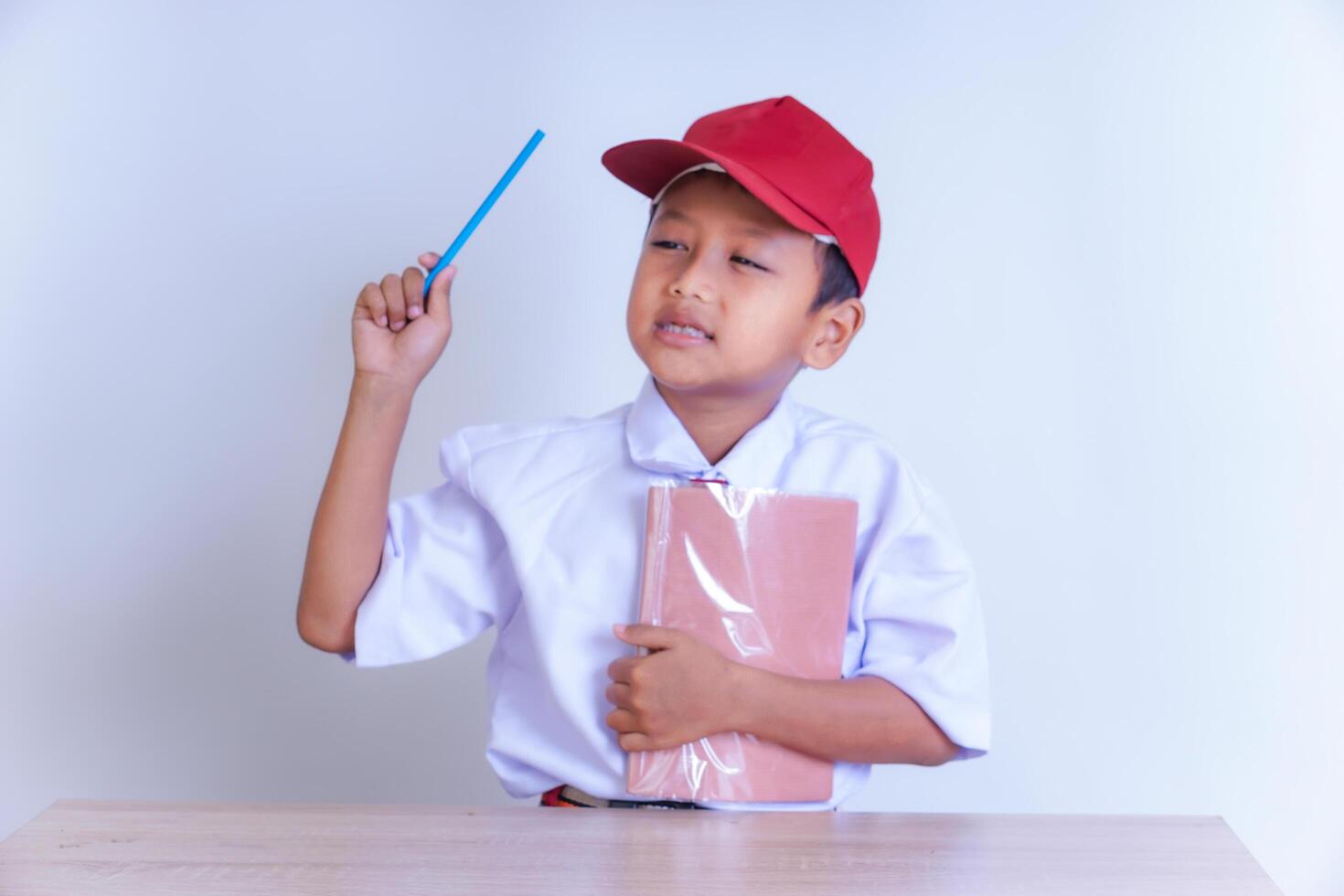 An Indonesian boy in school uniform, wearing a white shirt, tie and red cap, holds a blue pencil in his right hand while looking at her seriously. His left hand holds a book wrapped in plastic. photo