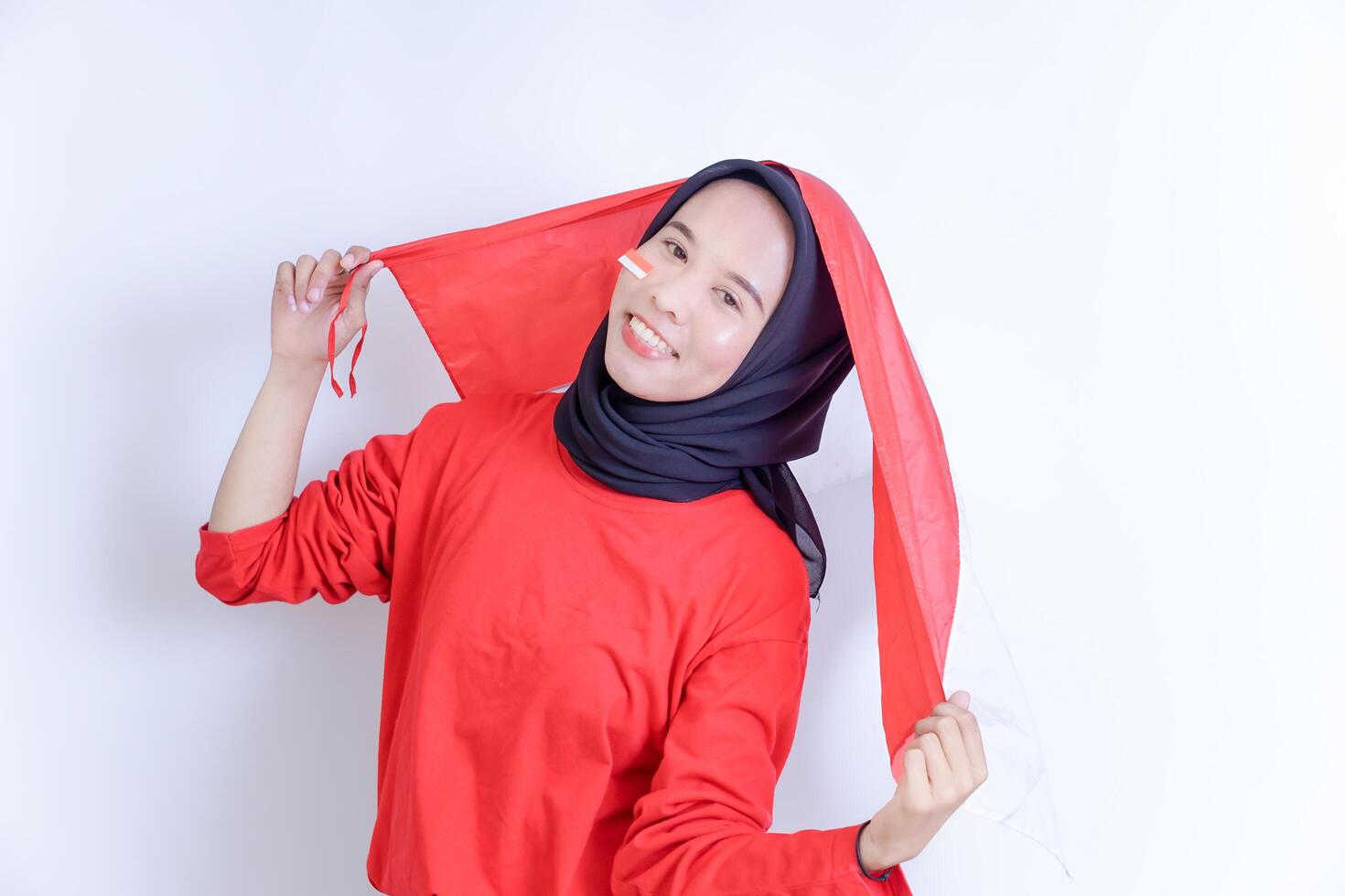 A Indonesian young woman wearing hijab is joyfully holding up the red and white national flag, her face adorned with a smile and a small flag sticker on her cheek, Isolated on white background photo