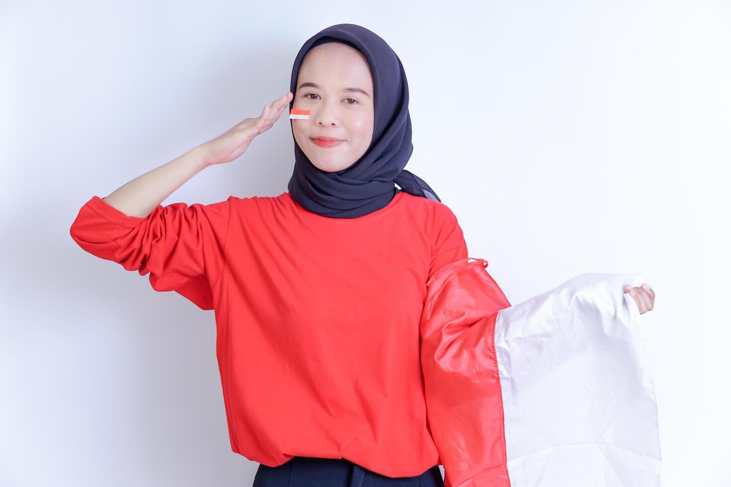 A young Indonesian woman wearing a hijab, she smiles warmly and salutes with her right hand, while holding a red and white flag on her left arm. celebrating Indonesia's independence day on August 17th photo