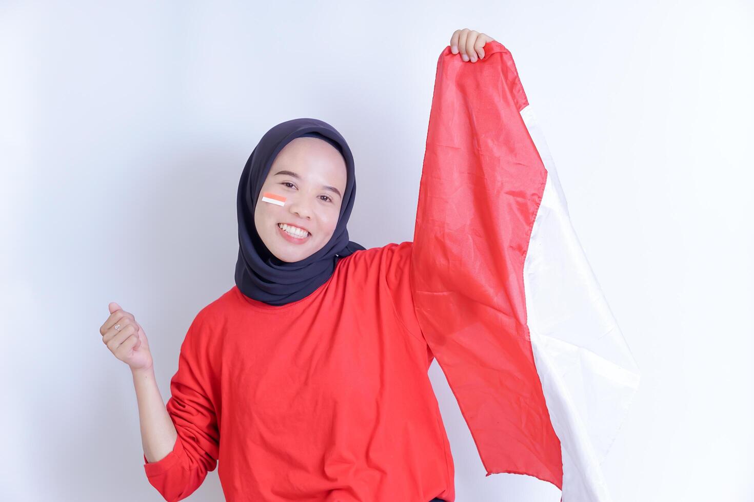 A young Indonesian woman wearing a hijab smiles happily while holding a red and white flag, with an Indonesian flag sticker on her cheek to celebrate Indonesia's independence day on August 17. photo