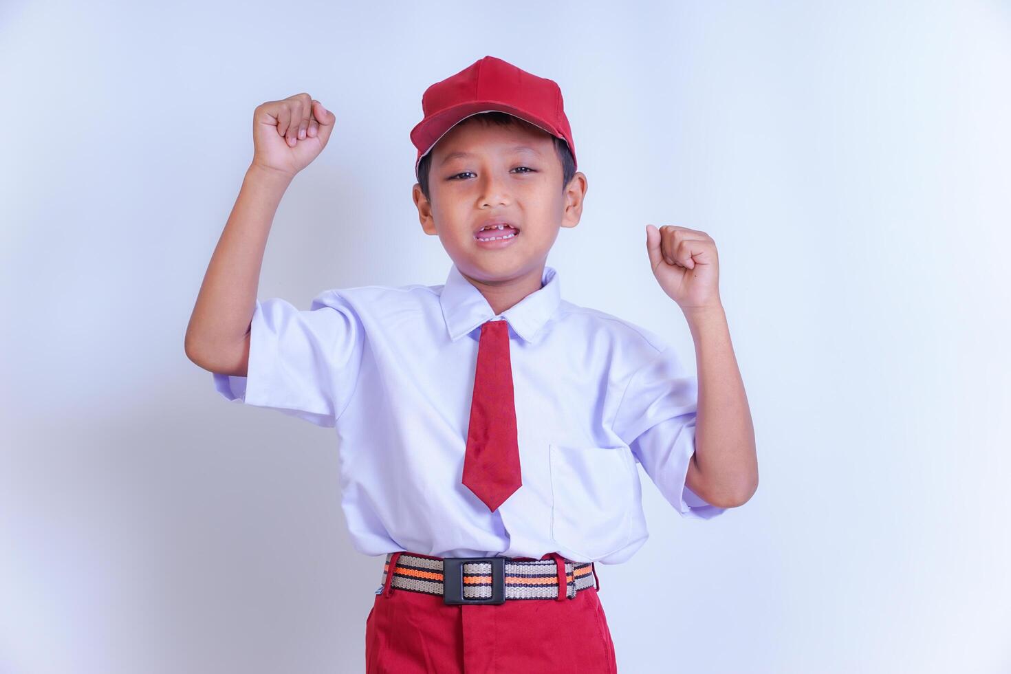 A Indonesian young boy in a school uniform poses confidently with both fists raised in the air. He is wearing a white shirt, red tie, and red cap. Isolated on white background photo