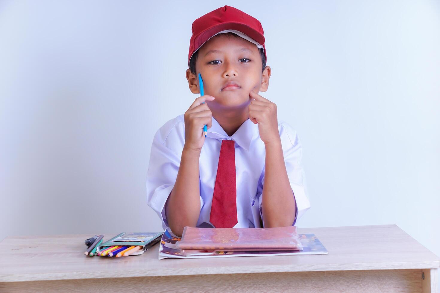 A Indonesian young boy in a school uniform sits thoughtfully with a pen, surrounded by study materials on a desk. photo