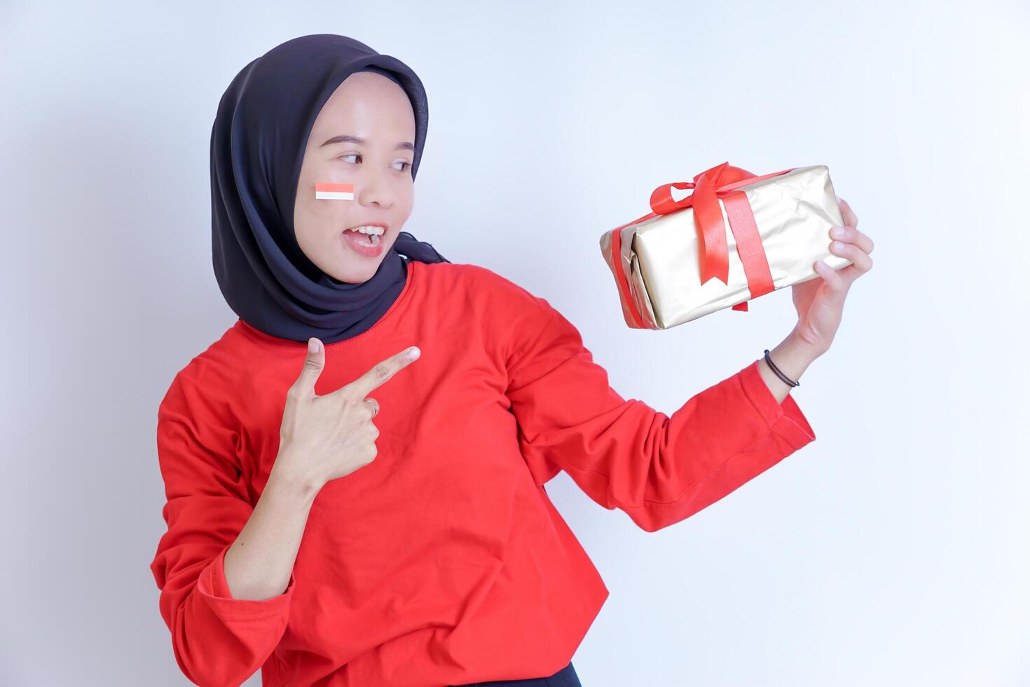 A smiling young Indonesian woman wearing a hijab, she holds out a gift wrapped in a red ribbon and points to it, drawing attention to the gift. celebrating Indonesia's independence day on August 17th photo