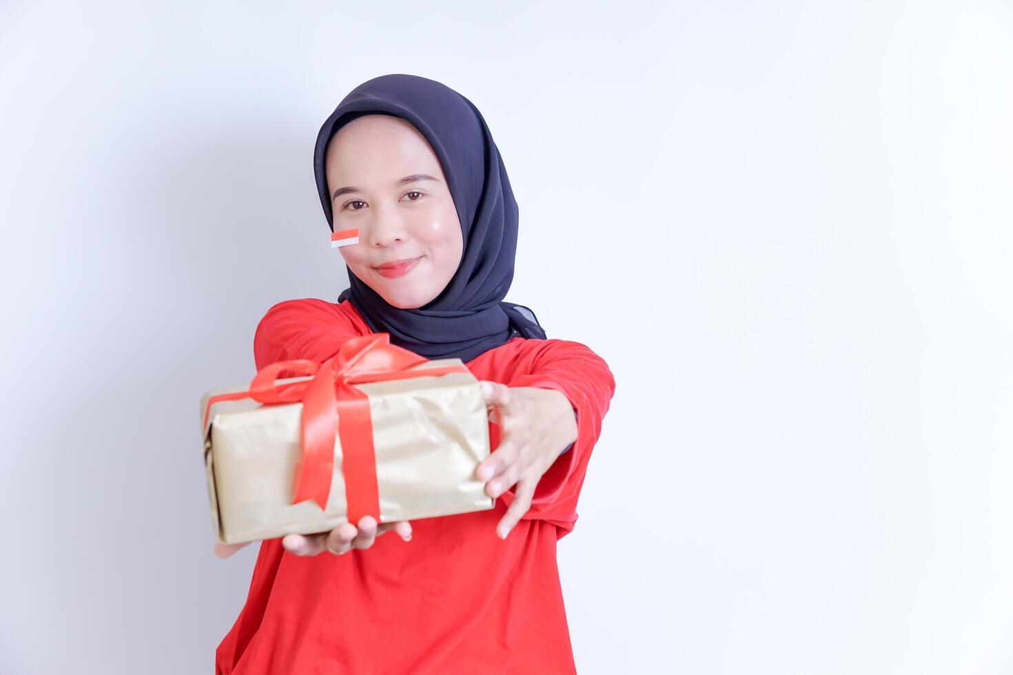 A young Indonesian woman wearing a hijab, with a small flag sticker on her cheek, smiling while giving a gift wrapped in a red ribbon, celebrating Indonesia's independence day on August 17. photo