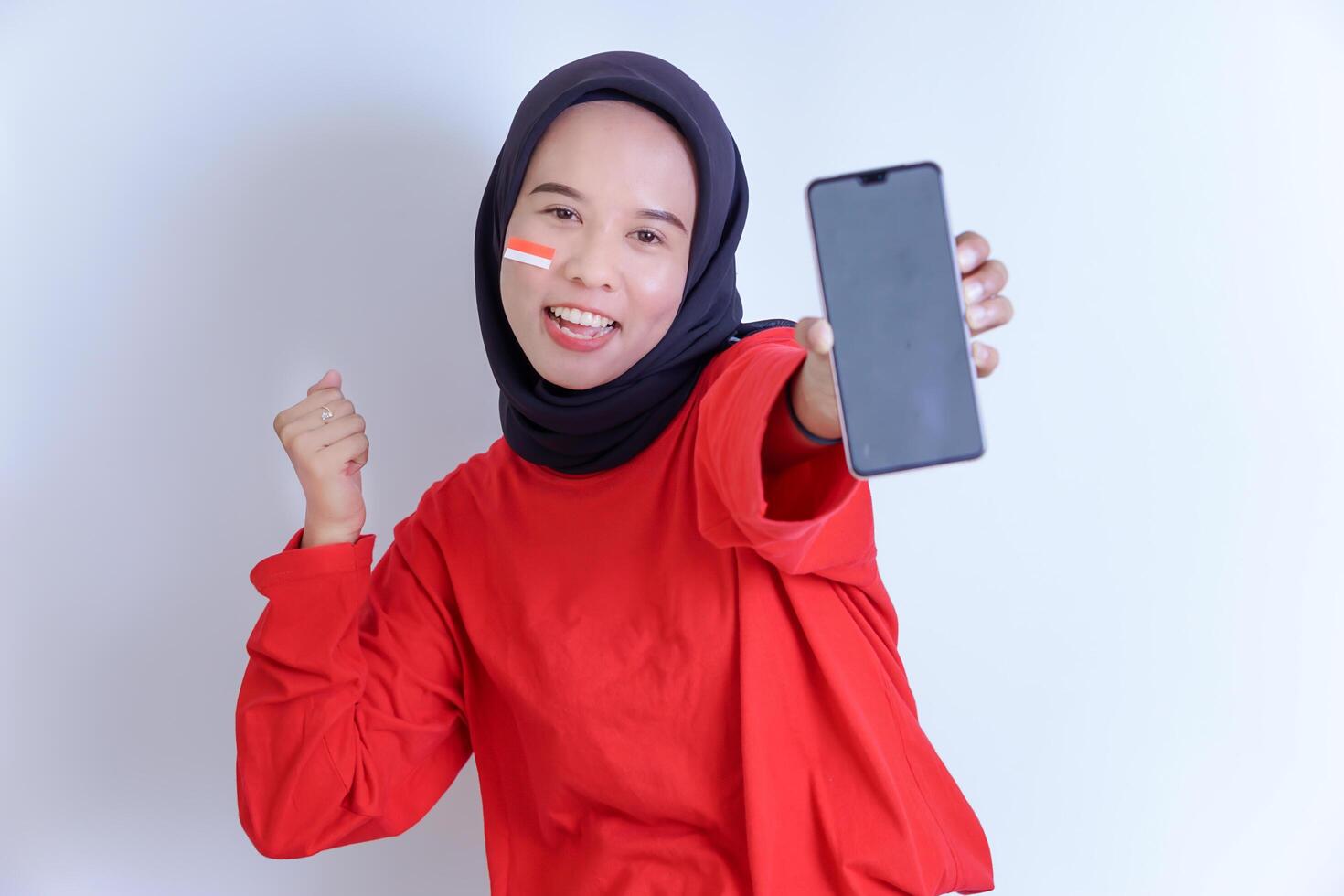 A young Indonesian woman wearing a hijab is smiling brightly while holding a phone and showing the national flag on her cheek, wearing a vibrant red shirt, isolated on white background photo