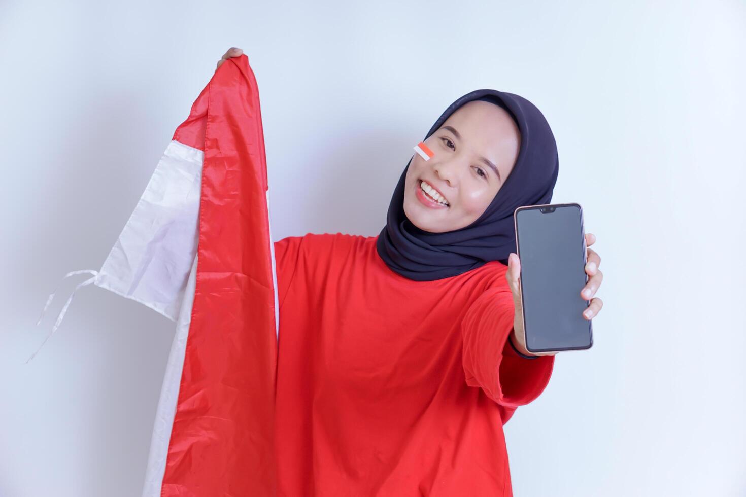 A young Indonesian woman wearing a hijab and a red shirt smiles brightly as she holds the Indonesian flag and proudly displays a smartphone, isolated on white background photo