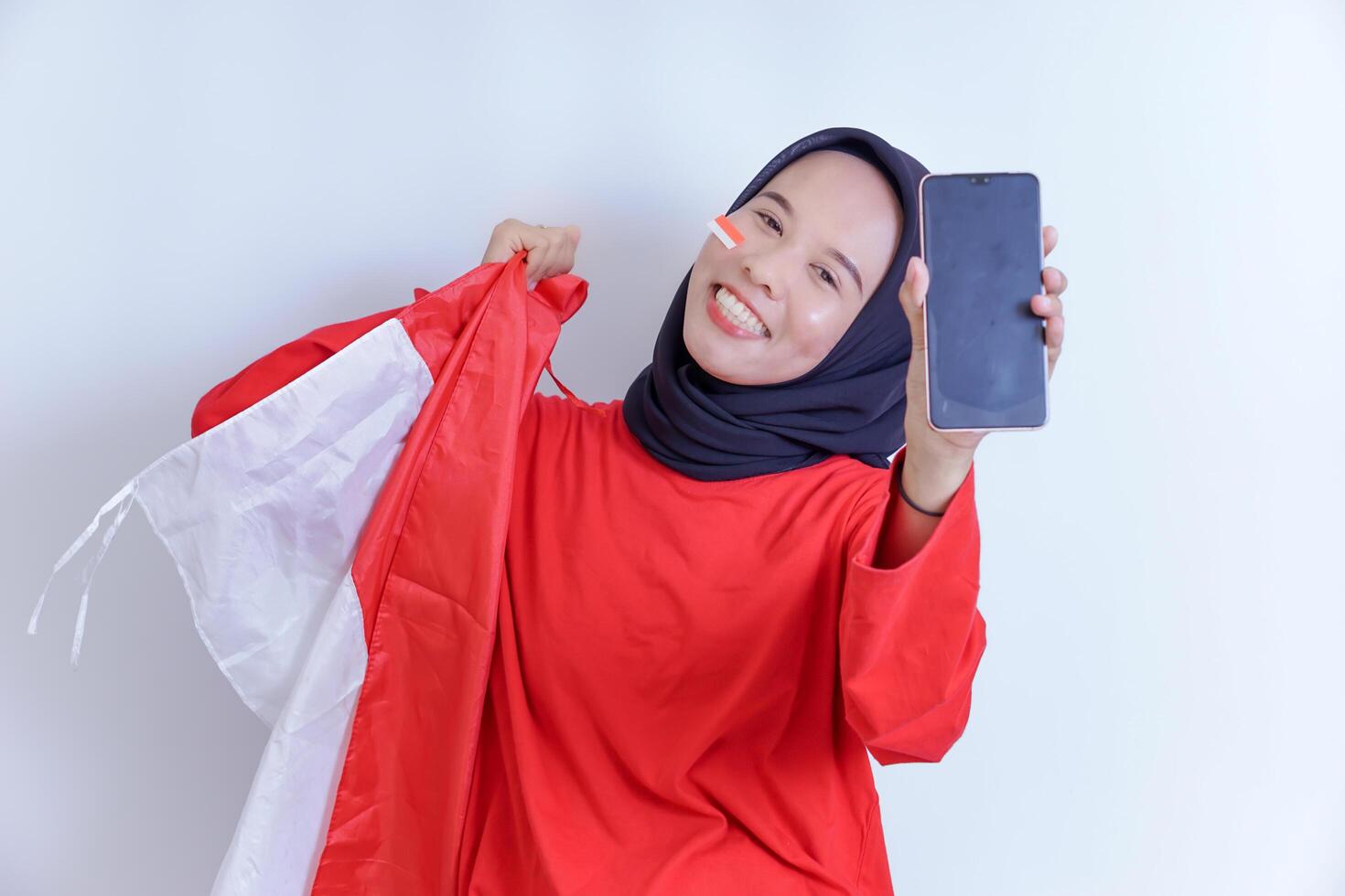 A young Indonesian woman wearing a hijab and a red shirt smiles brightly as she holds the Indonesian flag and proudly displays a smartphone, isolated on white background photo
