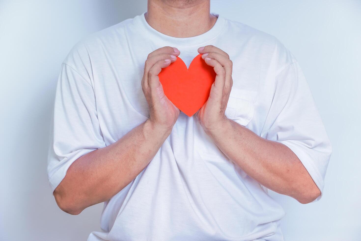 A person holds a red paper heart close to their chest, symbolizing love and affection against a light background. World Heart Day concept photo