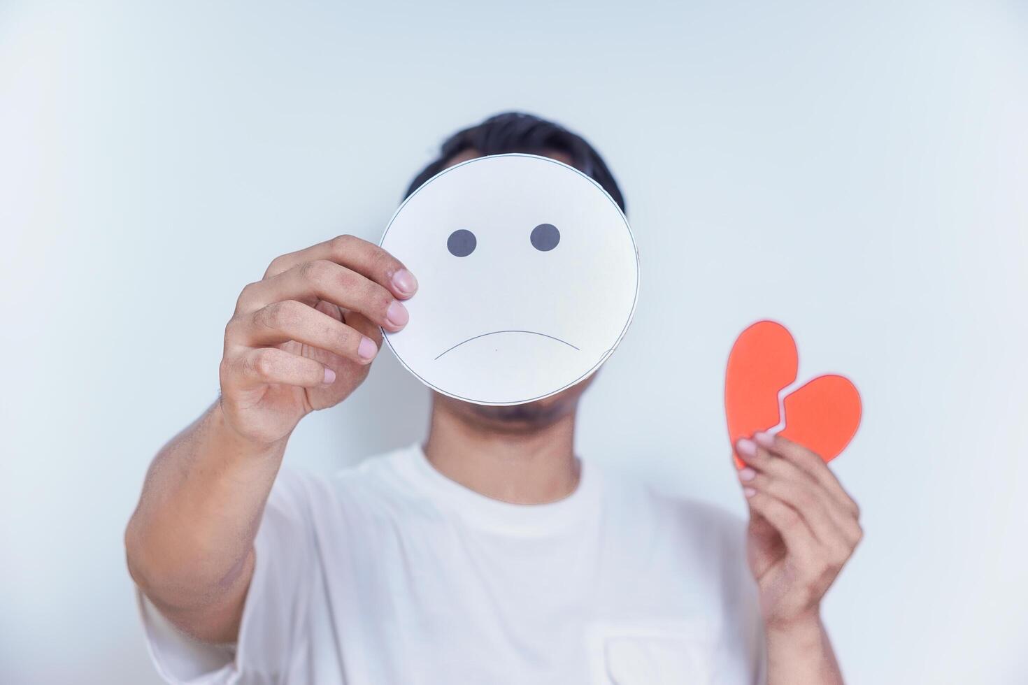 A person expresses sadness by holding a frowning face mask and a broken heart symbol, symbolizing emotional pain. world mental health day concept photo