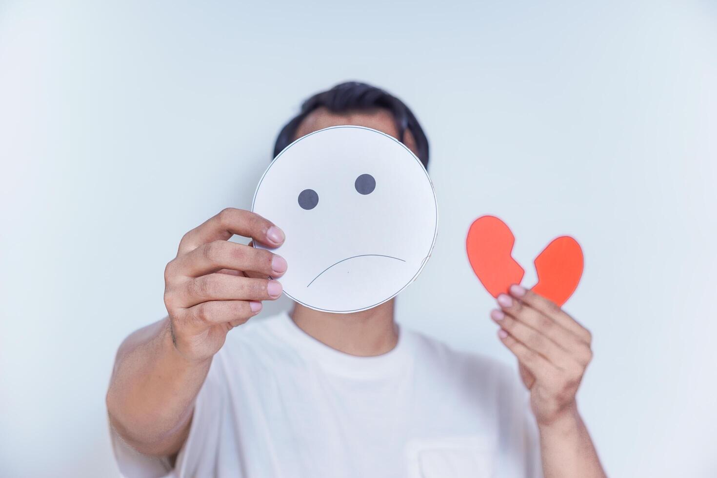 A person expresses sadness by holding a frowning face mask and a broken heart symbol, symbolizing emotional pain. world mental health day concept photo