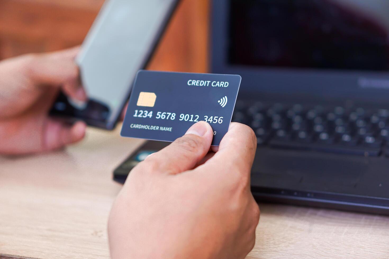 A person holding a credit card in one hand and a smartphone in the other while near a laptop. photo