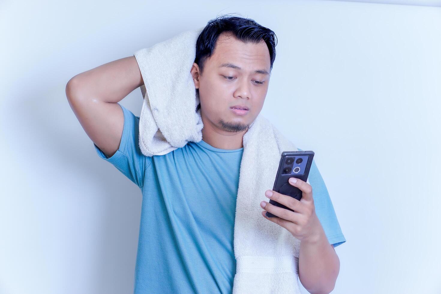 an Asian man with a disappointed expression while holding his smartphone and a towel on his shoulder photo