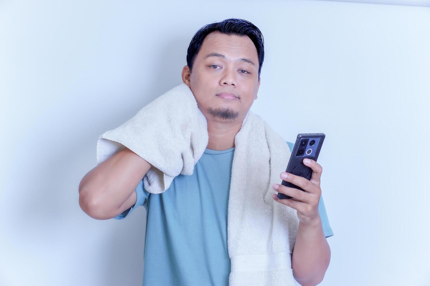 an Asian man with a disappointed expression while holding his smartphone and a towel on his shoulder photo