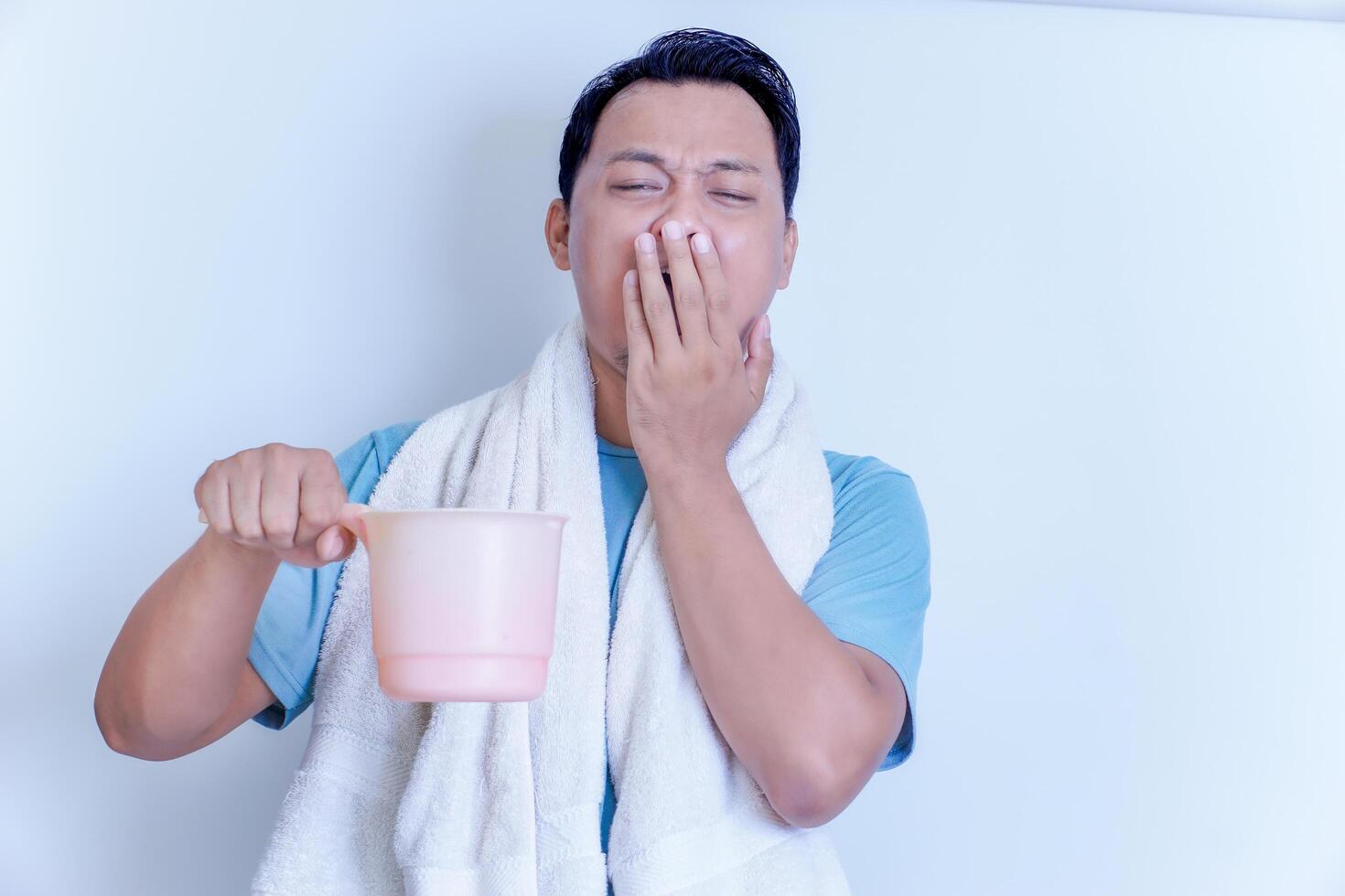 A Asian man yawns while holding a pink dipper, wrapped in a towel, looking sleepy. photo