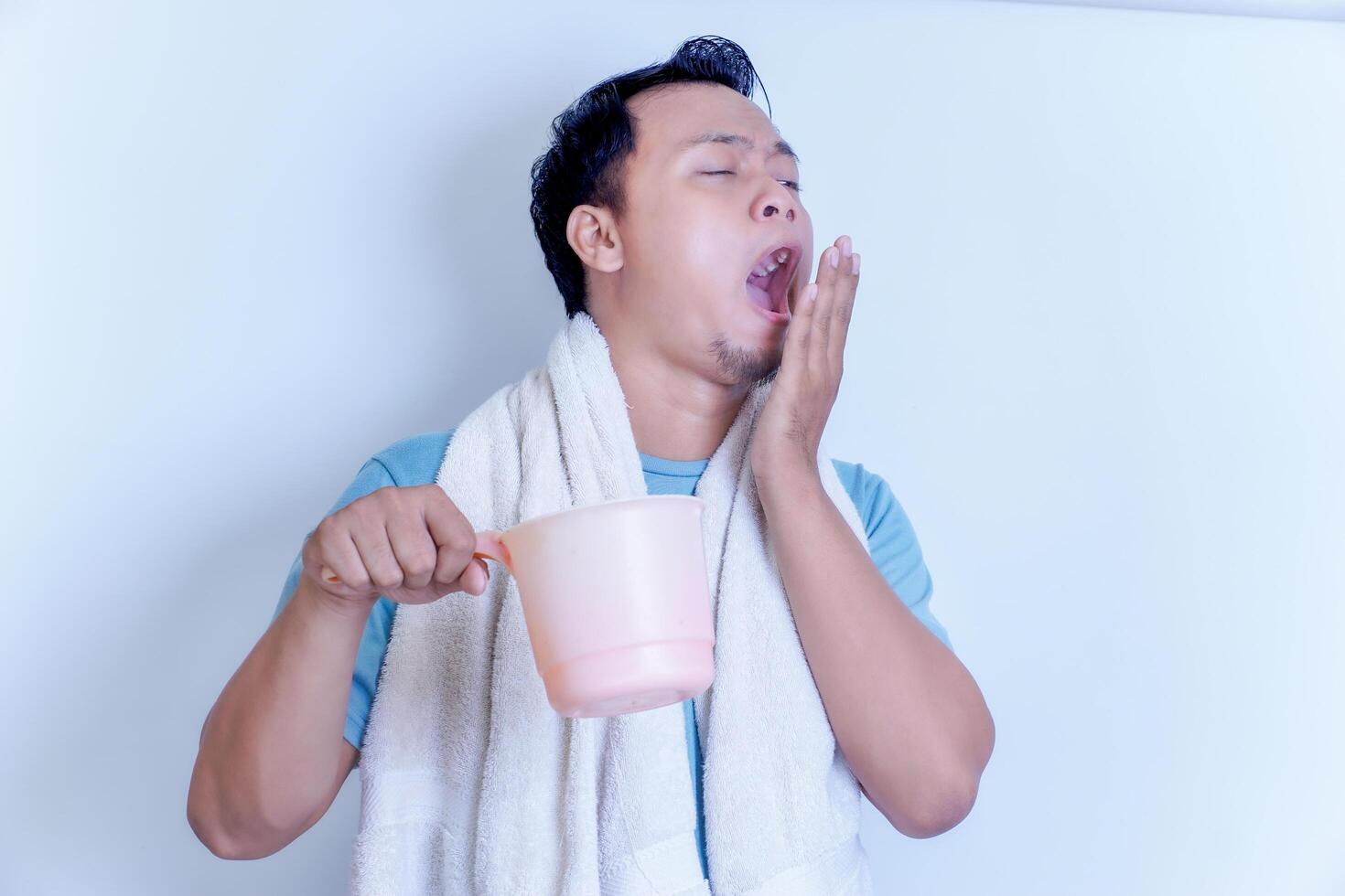 A Asian man yawns while holding a pink dipper, wrapped in a towel, looking sleepy. photo