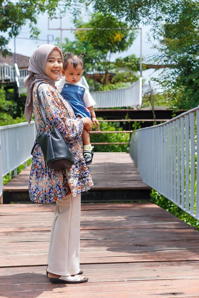 A happy young Asian muslim mom carries a child while walking on a wooden bridge surrounded by greenery. photo