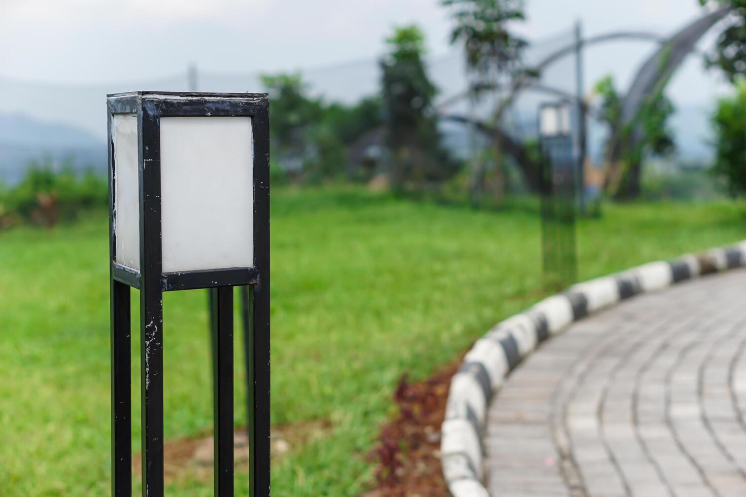 A modern lamp post stands in a lush green park, adding charm to the walking path around it. photo