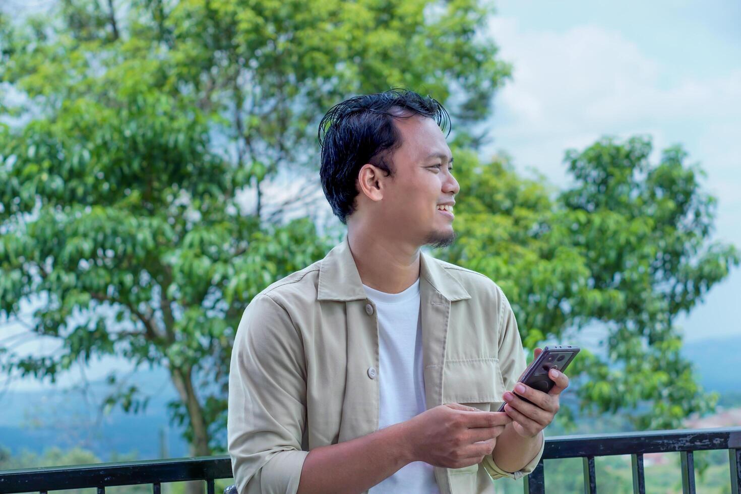 A asian man enjoys his time outdoors, smiling and using his phone amidst lush greenery. photo