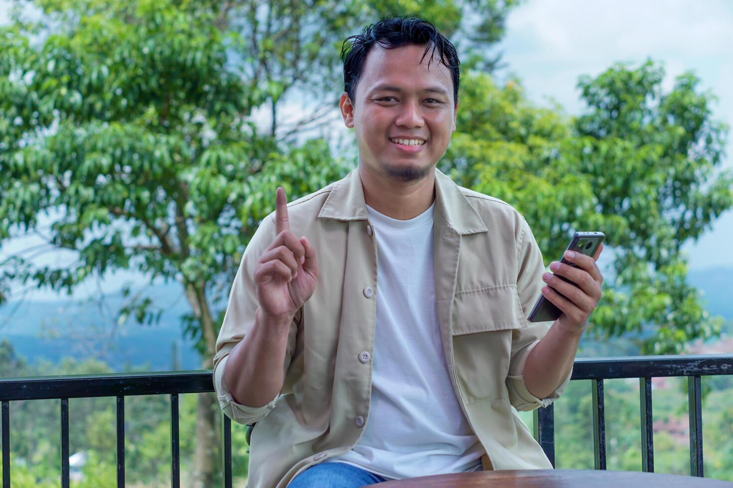 A asian man smiling and holding a phone, gesturing with one finger, against a scenic backdrop. photo