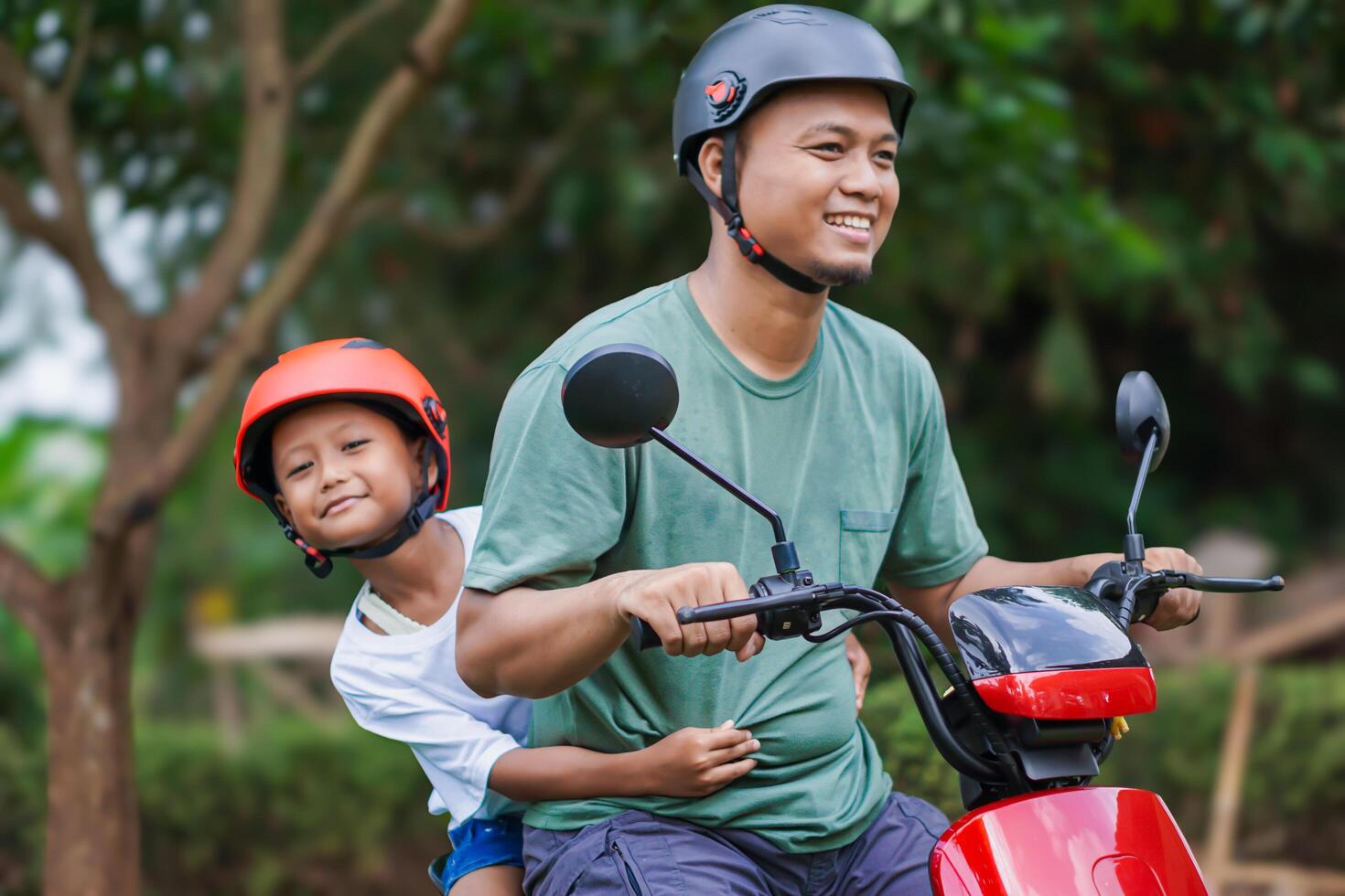 A asian father and son smiling together on a electric scooter, both wearing helmets in a green setting. photo