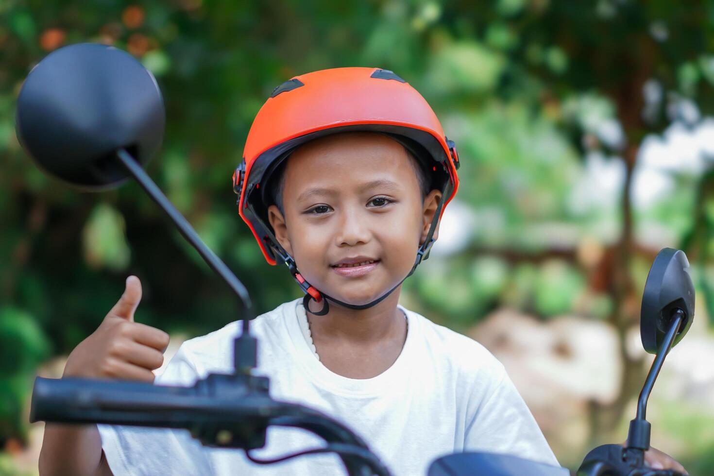 A young asian boy in a red helmet gives a thumbs up while sitting on a electric bike in outdoor photo