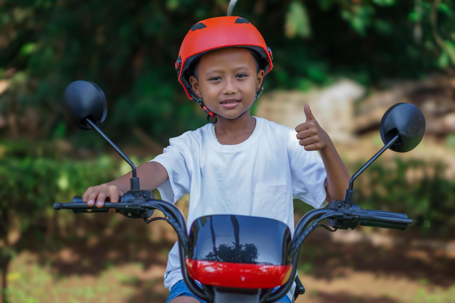 A young asian boy in a red helmet gives a thumbs up while sitting on a electric bike in outdoor photo